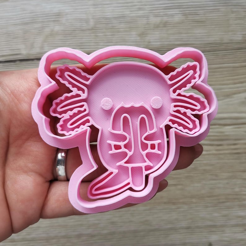 K&ouml;nnte beinhalten: Ein rosa Kunststoff-Ausstecher in Form eines Axolotls. Der Ausstecher hat die Form eines Axolotls mit detaillierten Kiemen, einem l&auml;chelnden Gesicht und einem abgerundeten Kopf. Der Ausstecher wird in einer Hand gehalten.