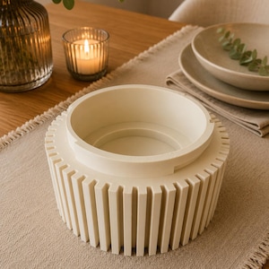 May include: A cream-colored cylindrical bowl featuring a series of vertical, evenly spaced slats around its exterior. The bowl has a smooth, raised inner rim and a flat base, presenting a structured, uniform design with a matte finish throughout.