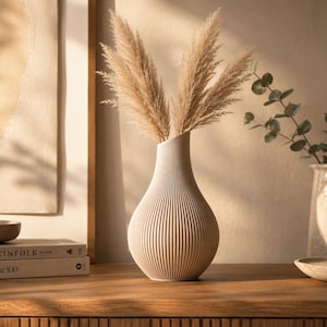 Könnte beinhalten: Eine beige Keramikvase mit vertikaler Rippenstruktur und verjüngtem Hals, gefüllt mit drei getrockneten Pampasgraswedeln. Die Vase steht neben einem Bücherstapel, einer kleinen Schale und einem zweiten Keramikkrug mit Eukalyptuszweigen.