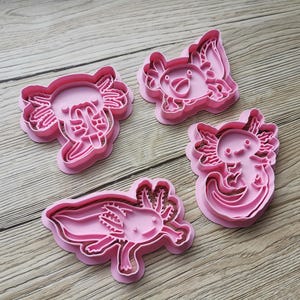 K&ouml;nnte beinhalten: Vier rosa Ausstecher in Axolotl-Form. Jeder Ausstecher zeigt ein anderes Axolotl-Design mit Details wie Kiemen, Augen und einem l&auml;chelnden Mund. Die Ausstecher sind aus Kunststoff und zum Backen bestimmt.