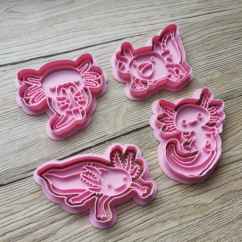 K&ouml;nnte beinhalten: Vier rosa Ausstecher in Axolotl-Form. Jeder Ausstecher zeigt ein anderes Axolotl-Design mit Details wie Kiemen, Augen und einem l&auml;chelnden Mund. Die Ausstecher sind aus Kunststoff und zum Backen bestimmt.