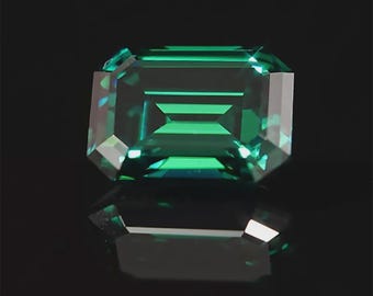May include: A faceted emerald-cut gemstone with a deep teal color. The gem is set against a black background, with its reflection visible below. The cut of the stone creates multiple facets that reflect light.