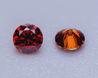 Puede incluir: Dos gemas redondas. Una es de color rojo intenso con muchas facetas, que reflejan la luz. La otra es de un color naranja más claro, también facetada. Ambas están sobre un fondo gris claro. Estas gemas podrían usarse en joyería.