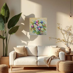 May include: A square floral painting featuring a vibrant bouquet of white, pink, blue, and purple flowers in a green vase. The artwork hangs on a beige wall above a white sofa with tan pillows. A large potted plant and a woven rug are also visible.