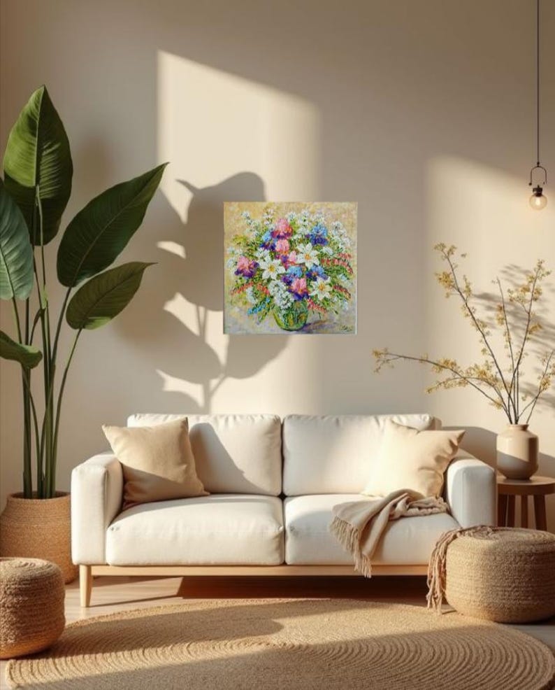 May include: A square floral painting featuring a vibrant bouquet of white, pink, blue, and purple flowers in a green vase. The artwork hangs on a beige wall above a white sofa with tan pillows. A large potted plant and a woven rug are also visible.