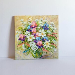 May include: Oil painting of a vibrant floral arrangement in a clear glass vase. The bouquet features white lilies, pink, purple, and blue irises, and small white flowers. The background is a textured, light yellow.