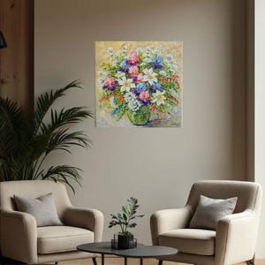 May include: An oil painting of a vibrant floral arrangement in a green glass vase. The bouquet features white, pink, purple, and blue flowers against a light yellow background. The painting hangs above two beige armchairs and a black coffee table.