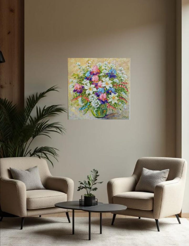 May include: An oil painting of a vibrant floral arrangement in a green glass vase. The bouquet features white, pink, purple, and blue flowers against a light yellow background. The painting hangs above two beige armchairs and a black coffee table.