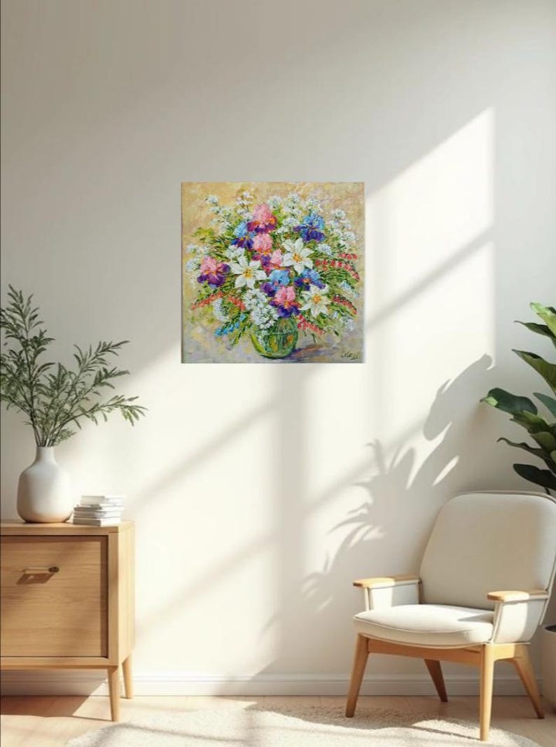 May include: An oil painting of a vibrant floral arrangement in a green glass vase. The bouquet features white, pink, purple, and blue flowers. The artwork is displayed on a beige wall, with a wooden cabinet and chair in the foreground.