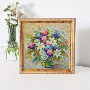May include: A framed floral oil painting featuring a vibrant bouquet of white, pink, purple, and blue flowers in a green vase. The painting has a gold frame and is set against a light background. A vase of white flowers is visible in the background.