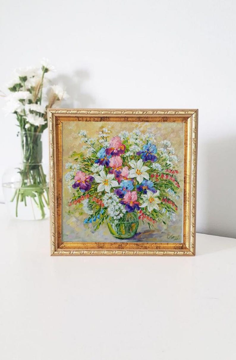 May include: A framed floral oil painting featuring a vibrant bouquet of white, pink, purple, and blue flowers in a green vase. The painting has a gold frame and is set against a light background. A vase of white flowers is visible in the background.