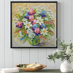 May include: A framed floral painting featuring a vibrant bouquet of flowers in a clear glass vase. The bouquet includes white lilies, purple and blue irises, and pink blooms. The painting is set against a light yellow background.