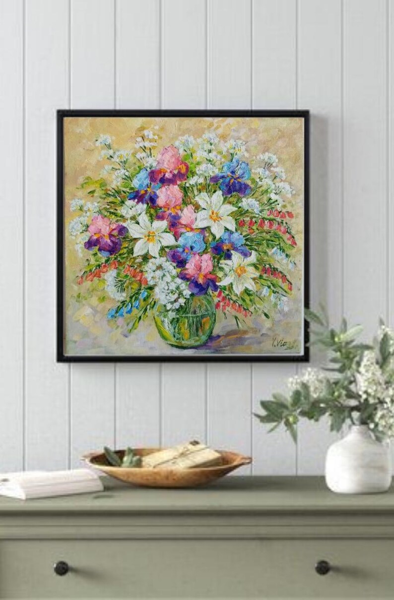 May include: A framed floral painting featuring a vibrant bouquet of flowers in a clear glass vase. The bouquet includes white lilies, purple and blue irises, and pink blooms. The painting is set against a light yellow background.