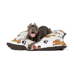 May include: A gray dog rests on a dog bed. The bed has a white top with a pattern of brown dogs and black paw prints. The bed has a brown border. The dog is smiling with its tongue out.