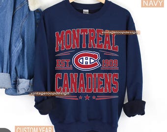 Puede incluir: Sudadera azul marino con el texto "MONTREAL CANADIENS" en rojo y blanco, el logotipo del equipo y "EST. 1909". La sudadera tiene cuello redondo y mangas largas con puños acanalados.