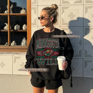 Könnte beinhalten: Schwarzes Sweatshirt mit dem Text "MINNESOTA WILD" in grünen und roten Buchstaben. Das Sweatshirt hat ein kreisförmiges Logo mit einem grünen und roten Design. Die Person auf dem Bild trägt schwarze Shorts und eine Sonnenbrille.