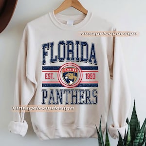 Puede incluir: Sudadera de color crema con el texto "FLORIDA PANTHERS" en azul marino, con una estética vintage desgastada. El diseño incluye un emblema circular con una cabeza de pantera y el texto "EST. 1993". La sudadera está colgada en una percha de madera.