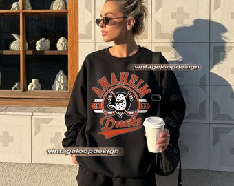 Może przedstawiać: Czarna bluza Anaheim Ducks z logo przedstawiającym maskę hokejową, skrzyżowane kije hokejowe i napisem "Anaheim Ducks 1993". Bluza jest połączona z czarnymi szortami. Obraz zawiera również tekst "vintageloopdesign".