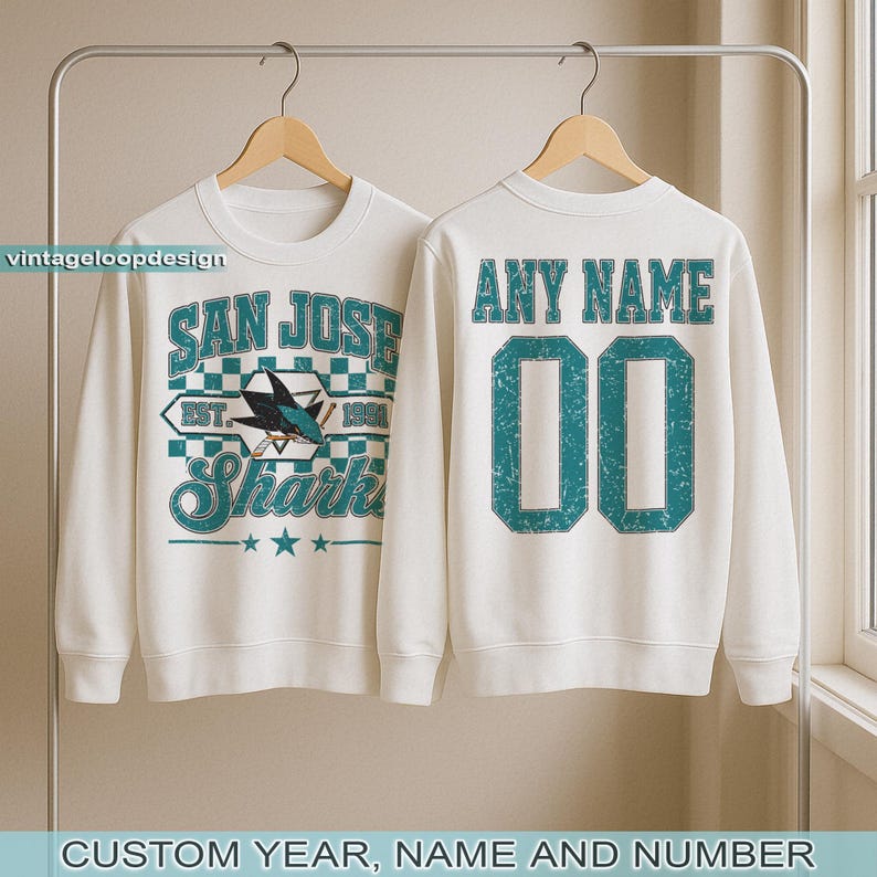 May include: White sweatshirt with teal graphics. The front features "San Jose Sharks" text and a shark logo. The back displays "Any Name" above the number "00". The design includes a vintage, distressed look.