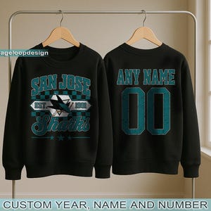 May include: Black sweatshirt featuring a "SAN JOSE Sharks" logo on the front and "ANY NAME" and number on the back. The logo includes a shark fin and the text "EST. 1991". The bottom of the image reads "CUSTOM YEAR, NAME AND NUMBER".