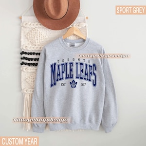 May include: Sport grey sweatshirt featuring the text "TORONTO MAPLE LEAFS" in blue, with a maple leaf logo and the year "1917". The sweatshirt is hanging on a wooden hanger.