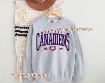 Puede incluir: Sudadera gris deportiva con el texto "MONTREAL CANADIENS" en letras azules y rojas. La sudadera tiene el texto "EST. 1909" y un logotipo. Un sombrero marrón cuelga encima.