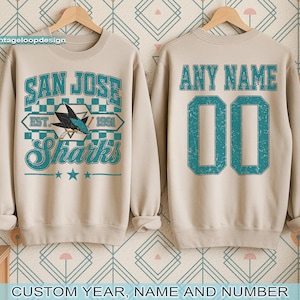 May include: Beige sweatshirt with "SAN JOSE Sharks" graphic in teal and white. The front features a shark logo and the text "EST. 1991". The back has "ANY NAME" and the number "00" in teal. The sweatshirt is displayed on a wooden hanger.