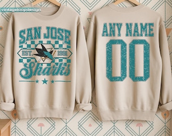 May include: Beige sweatshirt with "SAN JOSE Sharks" graphic in teal and white. The front features a shark logo and the text "EST. 1991". The back has "ANY NAME" and the number "00" in teal. The sweatshirt is displayed on a wooden hanger.