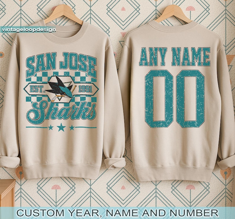 May include: Beige sweatshirt with "SAN JOSE Sharks" graphic in teal and white. The front features a shark logo and the text "EST. 1991". The back has "ANY NAME" and the number "00" in teal. The sweatshirt is displayed on a wooden hanger.