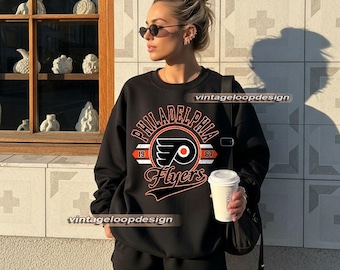 May include: Black crew neck sweatshirt with the text "PHILADELPHIA Flyers" in a circular design with the team logo. The sweatshirt also has the numbers "19" and "67". The person is wearing black shorts and sunglasses.