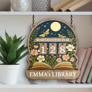 Puede incluir: Letrero de madera con un diseño floral y celestial. El letrero dice "BOOKS READ THIS YEAR" y muestra el número "128". La parte inferior del letrero dice "EMMA'S LIBRARY". El letrero está colgado en un estante con una planta y libros.