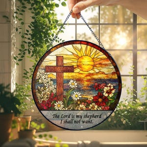 Op de afbeelding: Rond gebrandschilderd glazen raamkunst met een kruis, zon en bloemenontwerp. De tekst "The Lord is my shepherd I shall not want" is inbegrepen. Het kunstwerk wordt door een ketting vastgehouden.