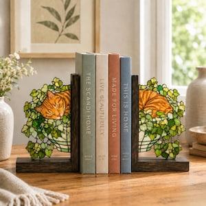 May include: A pair of dark wood bookends featuring stained glass panels with orange tabby cats sleeping in green ivy-covered planters. The bookends hold four books titled The Scandi Home, Live Beautifully, Made for Living, and This Is Home.