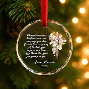 May include: A clear, round Christmas ornament with a red ribbon. The ornament features a wooden cross adorned with pink and white flowers. Text includes "Through patience, kindness, and care...Love, Emma 2025."