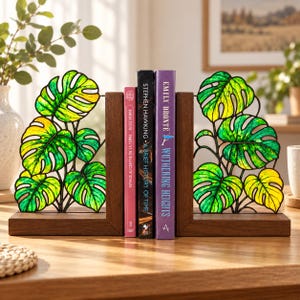 May include: A pair of bookends featuring dark wood bases and upright panels shaped like monstera leaves. The leaves are crafted with a stained glass effect in vibrant shades of green and yellow, held together by thin black metal frames.