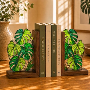 May include: A pair of L-shaped bookends made of dark wood, each featuring a stained glass panel shaped like a cluster of green monstera leaves. The bookends support four books with spines titled House Plants, The Kinfolk Home, Wild at Home, and Live Beautifully.