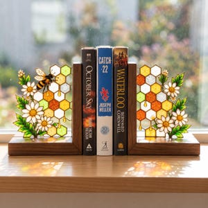 May include: A pair of wooden bookends featuring stained glass honeycomb patterns with white daisy flowers and a bee. The bookends hold three books titled October Sky, Catch-22, and Waterloo, displaying a colorful translucent design.
