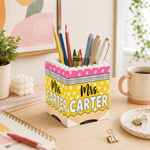 Puede incluir: Un portalápices cuadrado diseñado para parecer un lápiz amarillo con una goma de borrar rosa en la parte superior. El soporte presenta lunares blancos sobre un fondo amarillo y el texto Mrs. CARTER impreso en blanco y negro. Contiene lápices de colores.