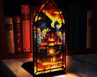May include: Gothic arch stained glass panel depicting a blue dragon and castle against a sunset. The artwork is set on a black base and is reminiscent of fantasy art. The panel is displayed in front of a bookshelf filled with books.
