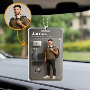 Op de afbeelding: Een auto-hangornament met de naam "James" en de titel "Programming Student". Het ornament toont een man met een koptelefoon en een rugzak, plus afbeeldingen van een laptop, koptelefoon, telefoon, notitieboek, toetsenbord, pen en muis.