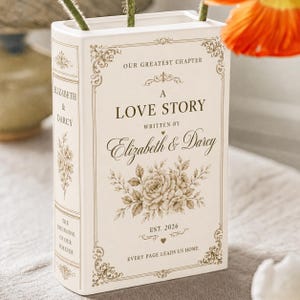 May include: A rectangular white ceramic vase shaped like a book with gold decorative borders and floral illustrations. The text reads: OUR GREATEST CHAPTER, A LOVE STORY, WRITTEN BY Elizabeth & Darcy, EST. 2026, EVERY PAGE LEADS US HOME.