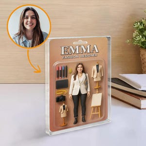 Op de afbeelding: Een modeontwerper actiefiguur genaamd Emma, verpakt in een doorzichtige plastic hoes. De figuur draagt een witte blazer en een zwarte broek. Accessoires zijn onder meer een camera, tablet en miniatuur kledingvormen. De verpakking leest "EMMA FASHION DESIGNER."