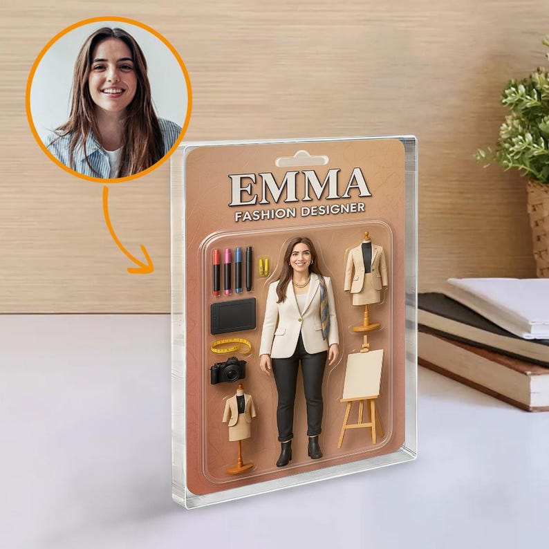 Op de afbeelding: Een modeontwerper actiefiguur genaamd Emma, verpakt in een doorzichtige plastic hoes. De figuur draagt een witte blazer en een zwarte broek. Accessoires zijn onder meer een camera, tablet en miniatuur kledingvormen. De verpakking leest "EMMA FASHION DESIGNER."