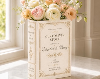 May include: A cream-coloured book-shaped vase featuring gold-toned decorative borders and text that reads A CHERISHED VOLUME OUR FOREVER STORY AUTHORED BY Elizabeth & Darcy EST. 04.12.2026 BOUND BY LOVE, WRITTEN IN TIME, filled with pastel flowers.