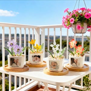 May include: Four small white square planters with glass flower sculptures and photos. Each planter sits on a round wooden coaster. The flowers are purple, yellow, white, and orange. The planters have the text "I love you, Mom" and "I love you, Grandma".