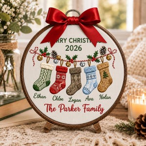 May include: A circular wooden embroidery hoop frame containing white fabric with a printed design of five hanging Christmas stockings in green, red, brown, blue, and tan. The text reads Merry Christmas 2026, The Parker Family, and five names.