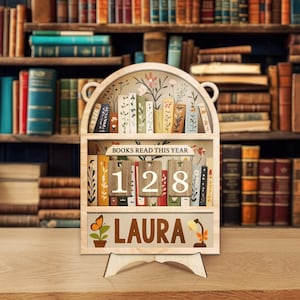 May include: A wooden bookshelf-shaped display with the text "BOOKS READ THIS YEAR" and the number "128". The name "LAURA" is displayed below, along with a butterfly and a lamp illustration. The background shows a library.