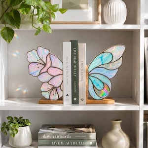 May include: A pair of stained glass bookends featuring iridescent butterfly wings in pastel pink and blue tones. Each wing is mounted on a rectangular wooden base, designed to hold books upright with a delicate, shimmering finish and dark metal lines.