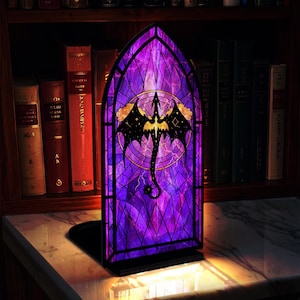 May include: A stained glass-style bookend featuring a dragon design. The bookend has a purple and black color scheme with a dragon silhouette in the center. The dragon is set against a background of geometric shapes and a night sky.