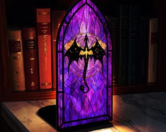May include: A decorative bookend with a stained glass effect, depicting a dragon. The design features a purple and black colour palette, with a dragon silhouette at the centre. The dragon is set against a geometric and night sky background.